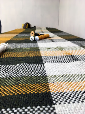 A low-angle shot of green, yellow, and white plaid fabric spread flat on a table, with yellow-white plastic bobbins holding matching yarn stacked about halfway down the table. From left, lengthwise marigold, dark green, white, and dark green stripes show, with crosswise strips of the same colors plus light green.