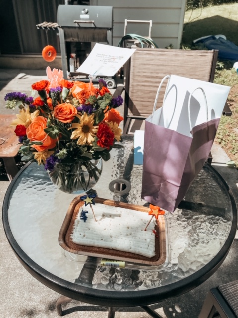 a table filled with birthday gifts including a small cake with two candles, a bouquet of flowers, and a purple gift bag 