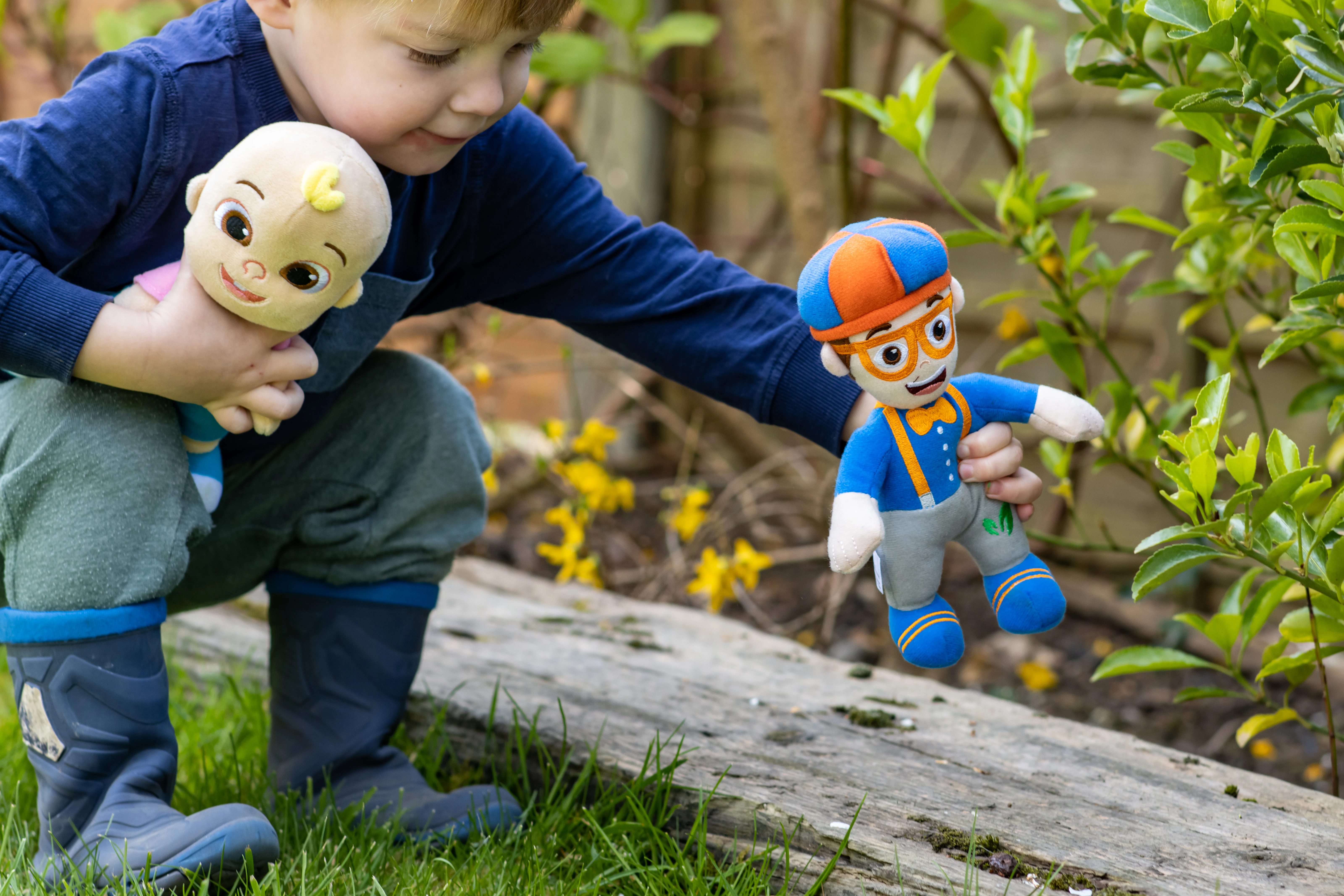 A preschooler in the garden with two plush toys: Blippi and JJ from CoComelon