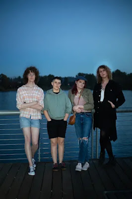 Four trans people standing in front of a waterfront, leaning against a white steel railing, looking at the camera.