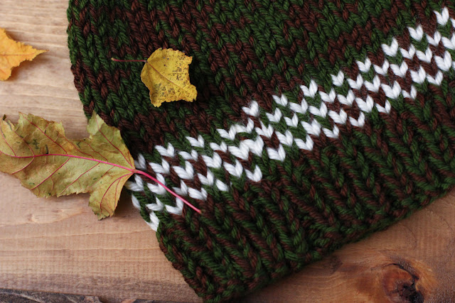 Close-up near brim of flat lay of a green and brown knitted hat with cream fair isle pattern accents lying on a wood surface with dried autumnal leaves.