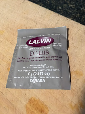 A grey packet of champagne and sparkling wine yeast, with the "Lalvin" label and other writing in white and mauve, on a pale wooden cutting board.