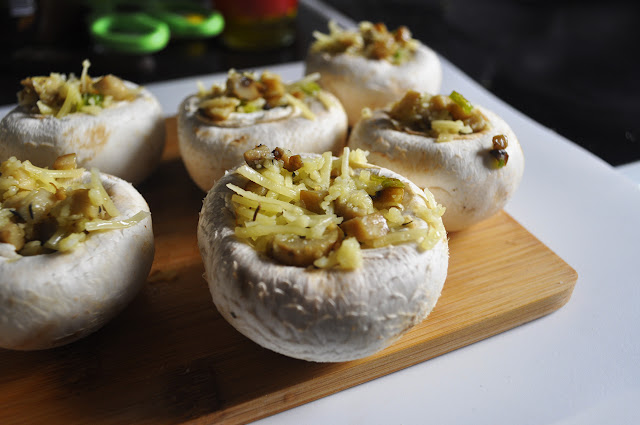 en una mesa de cocina, sobre unatabla de madera hay 6 champiñones blancos grandes rellenos, se ven las tiras de queso.