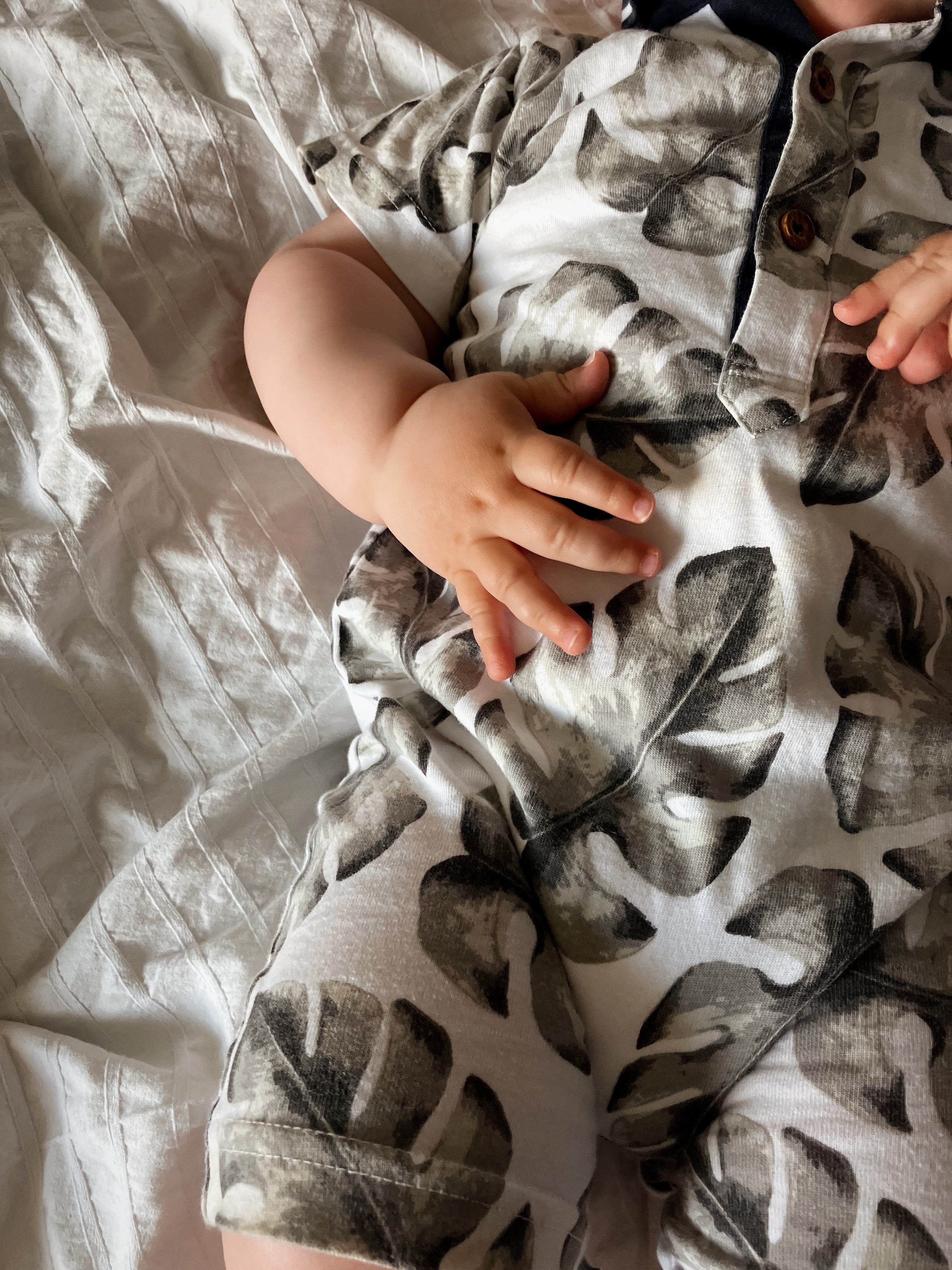 a baby lying on a white bedsheet wearing a romper patterned with monstera leaves. You can only see the romper and the baby's hands