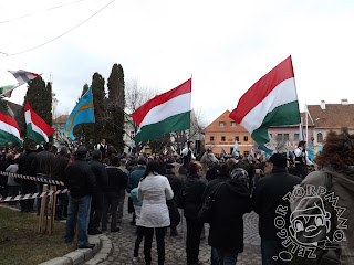 Szöveg: ...lovukat peckesen megülő huszárokat és válogatott cigánylegényeket... Kép: Emberek sorfala között óriási magyar és székely zászlót lobogtatva vonul be egy sor fiatal egyes sorban.