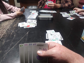 A game of Antidote in progress. Three players can be seen across the table from the point of view of the current player, who's holding his cards up where they can be seen. The current player's hand consists of a light green 4, a pink 2, a blue 2, and a purple 2. Several piles of cards can be seen in front of each player. The box lid is face down in the centre of the table, with an additional card in it.