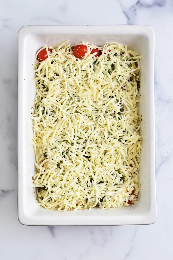 A casserole dish with foil removed, revealing the pesto-coated chicken topped with a generous amount of shredded mozzarella cheese, ready to be placed back in the oven to melt the cheese, in the process of making mouthwatering baked pesto chicken.
