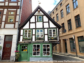 A small sized two-storey timbered house with a bright green entrance door and colourful windows and a wooden sculpture of a man in its gable.