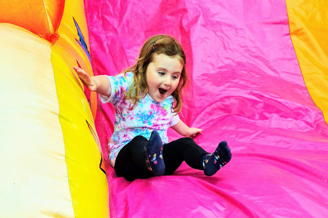 Image of a young girl wearing a tie dye t shirt and black leggings sliding down a bright pink and yellow inflatable slide.