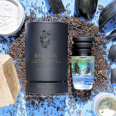 The bottle and black box of Russian Tea by Masque Milano lying flat on a bed of black tea, next to two face cream jars, rubber pieces and a homemade soap bar