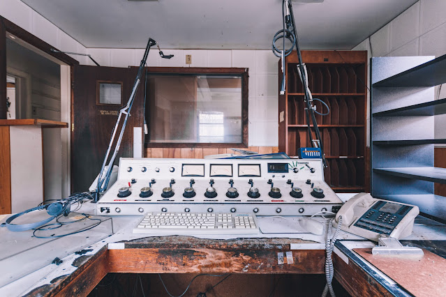 A deserted radio broadcast desk, with a vintage audio mixer, microphone stand, and telephone, all coated in dust. An empty rack that once held CDs or tapes looms in the background, with a window overlooking another abandoned room, reflecting the silence that has replaced the once lively sound of broadcasting.