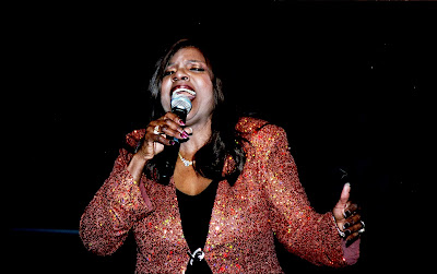 Gloria Gaynor singing wearing a gold and red jacket against a black background