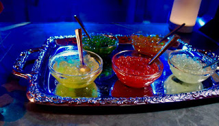 Semicircular glass clear bowls containing red, yellow and white small spherical balls of boba on a silver rectangular table with a blue cloth on a dark background 