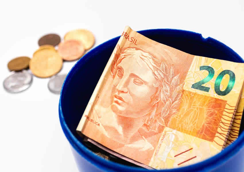 Imagem com fundo branco onde mostra um recipiente azul com uma nota de 20 reais dentro e algumas moedas ao lado.