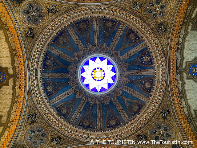 A rose window with white and blue decoration in the golden decorated ceiling of a synagogue.