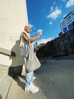 Photo taken from a low angle of a young male wearing a trendy outfit- blue jeans, thick white trainers, white t-shirt and a large bag on his arm. He is holding his phone out at level with his chest.
