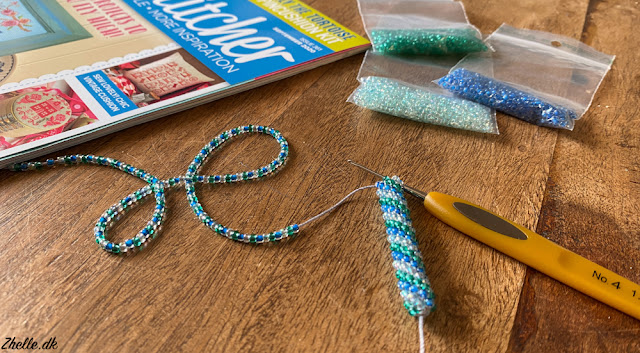 a crocheted blue green and white bead rope in the making with bags of beads in the background and a CrossStitcher magazine