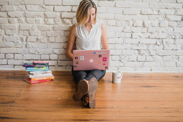 jovem mulher sentada em um chão aparentemente de madeira com as pernas cruzadas usando um laptop cinza aberto. Ela está encostada em uma parede de tijolos brancos e ao seu lado há uma pilha pequena de livros.