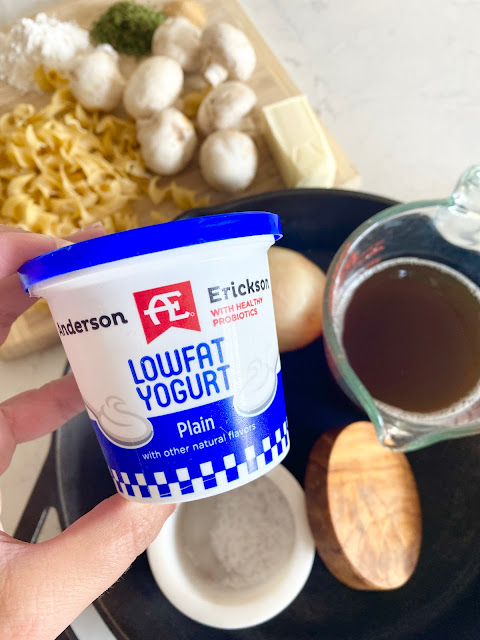 A hand holding AE Dairy plain yogurt in front of other beef stroganoff ingredients.