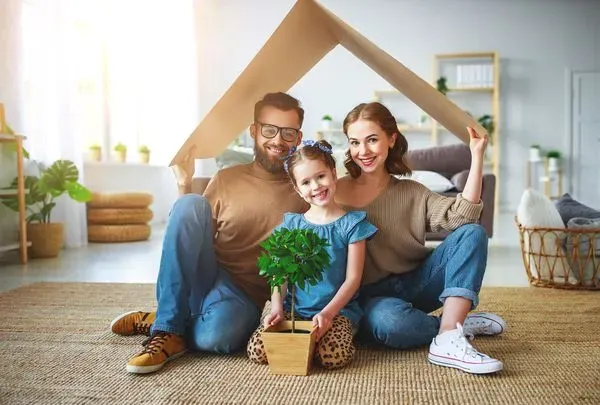 Família composta por pai, mãe e uma filha reunida debaixo de uma caixa de papelão em formato de casa representando a união dentro de casa.