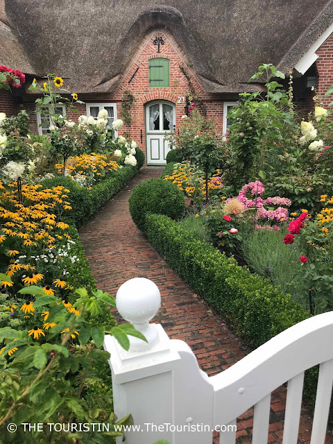 Path through a cottage garden leading to the white and green wooden door of a red brick cottage..