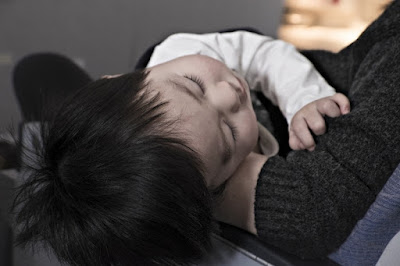 Young tot with black hair, being held on the lap of an adult