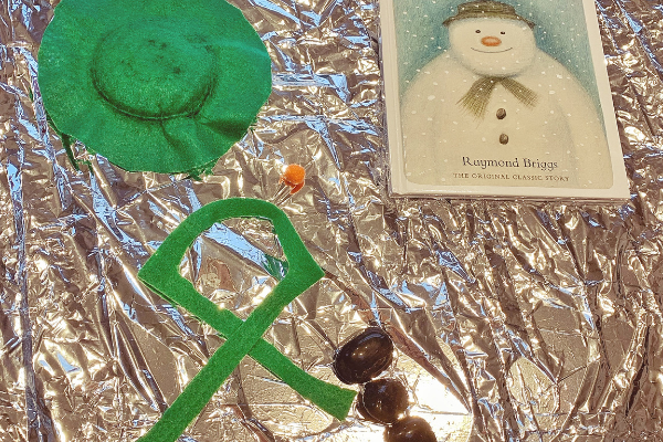 The Snowman book by Raymond Briggs with green felt hat and scarf and black pebble buttons on a tuff tray