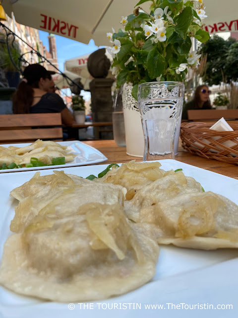 Two plates of Polish pierogi, a glass of water and white blooming flowers in a white pot on a wooden table in an outdoor restaurant.