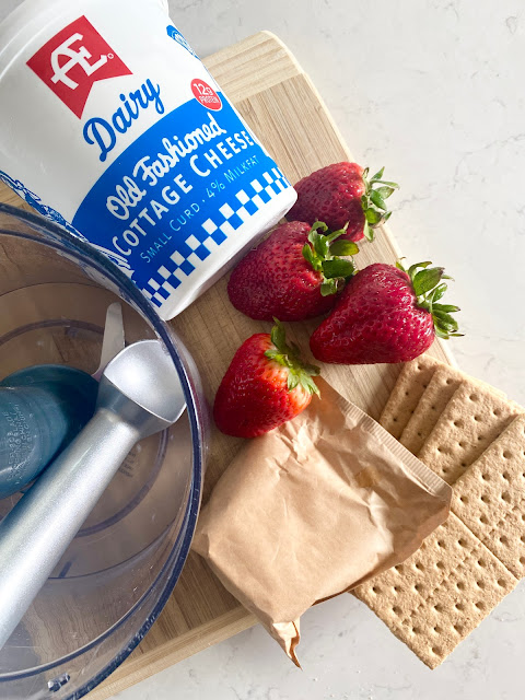 Food processor, strawberries, ice cream scoop, graham crackers and pudding mix sitting on a wooden cutting board.