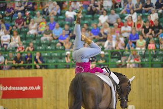 A disabled person balancing upside down on a dark bay horse in front of an audience in a large indoor arena