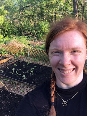 Selfie of a smiling, slightly sunburnt redheaded woman wearing a black mock-turtleneck and gold necklace with a ring and centered white stone pendant. Behind her is a raised bed with dark, wet earth and a neat block of flopped-over tomato plants, enclosed by mulched paths and a low lattice fence. Brush and trees fill the background.