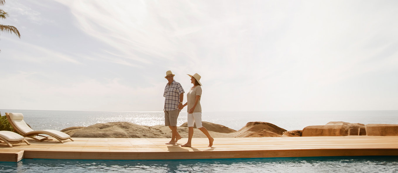 Photo of two people holding hands walking alongside a pool with a sunny background behind them which also features the sea and various rocks.