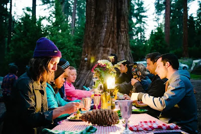 People gathered around a table under a tree. There is a big pine cone in the forefront.