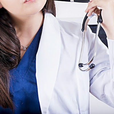 A nurse in blue scrubs and white medical jacket removing stethoscope from neck.