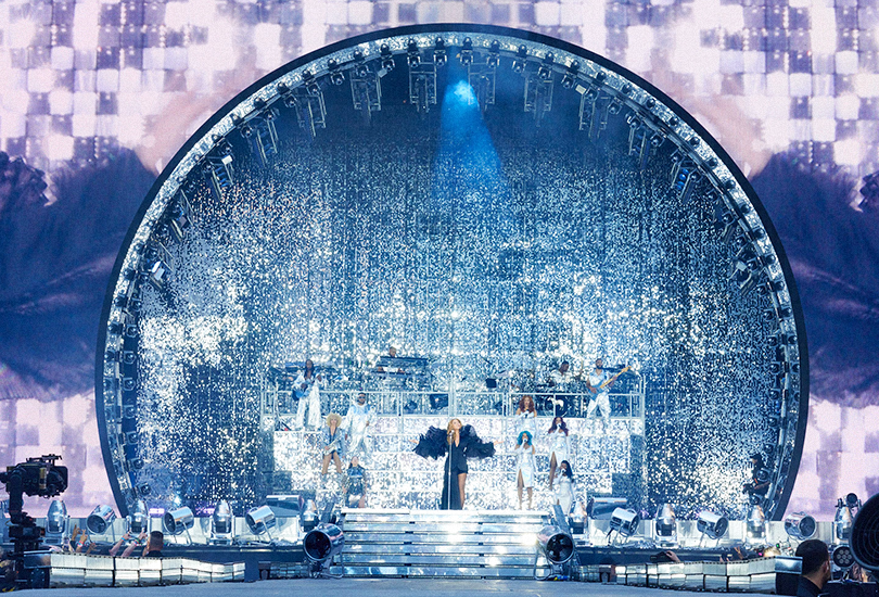 A shot of Beyoncé performing on the Renaissance World Tour. Beyoncé is stood in front of the giant circular aperture of the LED wall, backed by her band, backup singers, all stood in front of a backdrop of shimmering silver.