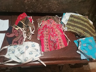 A dozen patterned fabric facemasks with elastic or self-fabric ties, and piles of cut fabric for additional masks. 