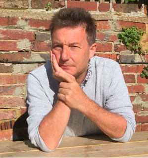 Rex Burke, a white man with brownish hair sitting leaning on a wooden table inn front of an orange brick wall.