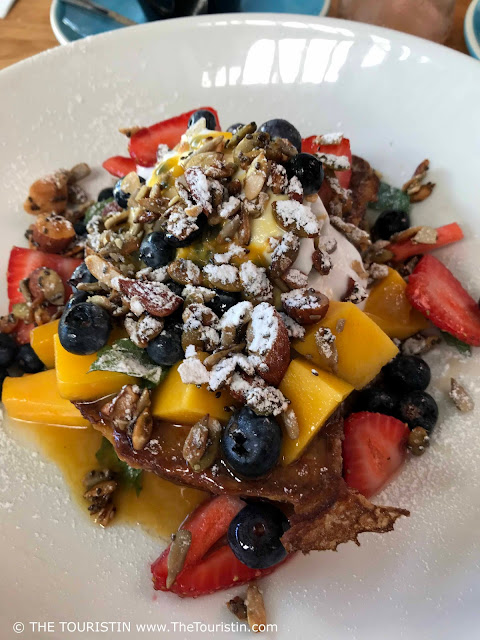 French toast, decorated with blueberries, sliced strawberries, diced mango and nuts, served on a white plate.