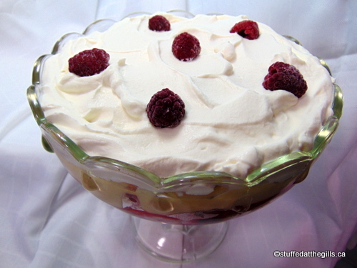 Newfoundland Fruit & Jelly Trifle layered in a clear footed trifle dish. Top layer cream garnished with raspberries, next layer bananas, then custard, fruit and jelly and last layer cake.