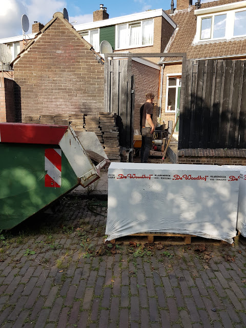Tegen de achtergevel van een schuurtje bij een achtertuin staan oude stoeptegels opgestapeld. Op de straat staat een grote puincontainer. Ernaast staan nog ingesealde nieuwe tegels van Woudhof uit Klarenbeek.
