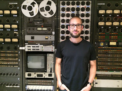 Image description: man posing in front of old-fashioned gray communications gear. There are analog meters, a tape-to-tape reel, a cathode-ray tube screen surrounded by switches, buttons, and dials. Directly behind the man is a plugboard with dozens of sockets.