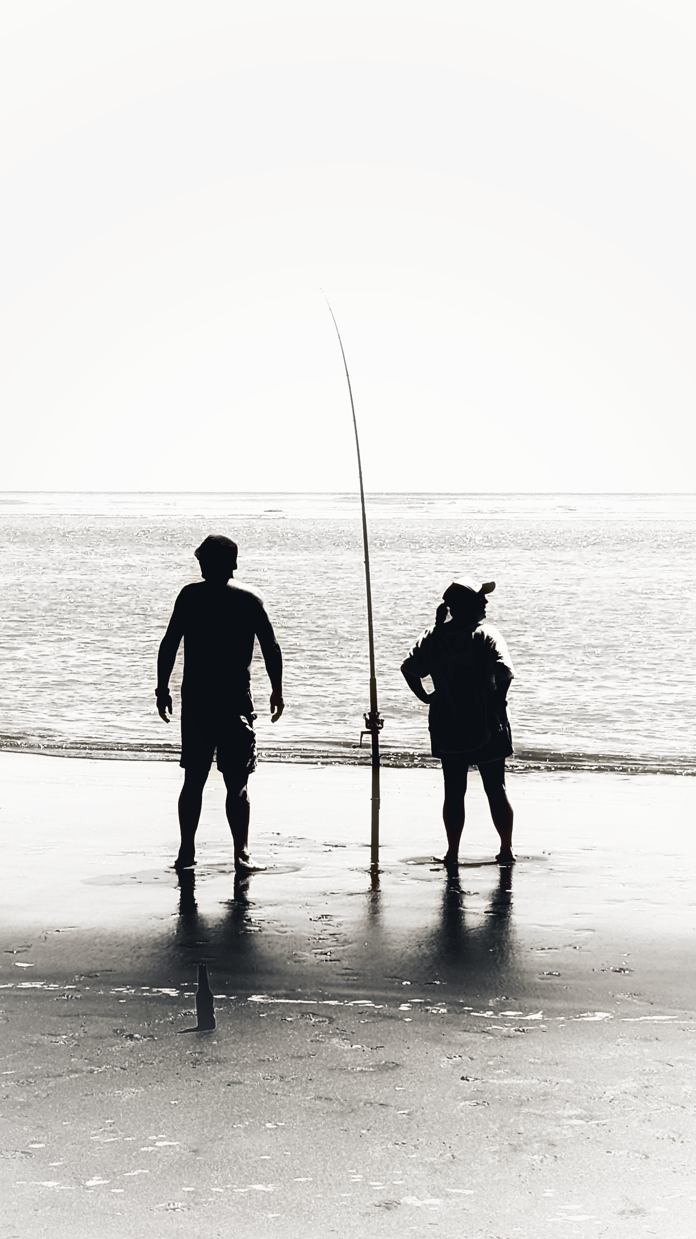 Stark black and white photo (high contrast) of two figures looking opposite ways down the beach as they fish, a fishing line stands hopeful in the sand between them