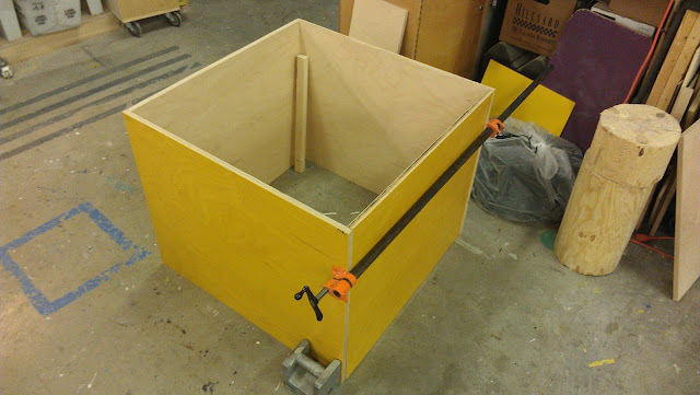 A large yellow wooden box of 34" x 34" x 34" sits on the ground with a large black clamp and a silver square weight holding it together.  There is no top or bottom yet to the box so a thin piece of wood about two feet long can be seen in the box leaning against the side.  The floor is a grey concrete and there are paint splatters on the floor.  There are cardboard boxes behind the wooden box and a sanded down wooden post as well.