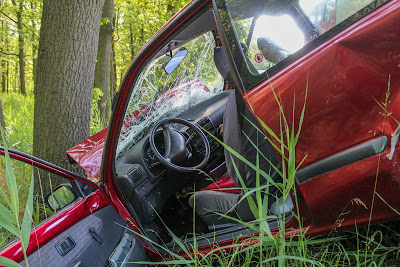 Red Car with busted glass open door leaning on tree
