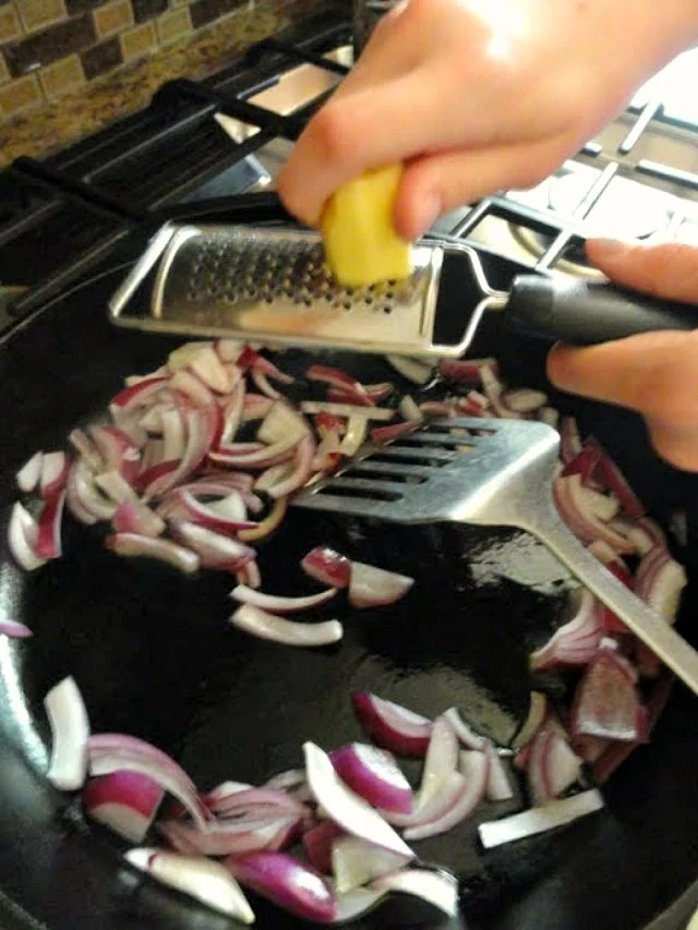 A cast iron pan with sautéed red onion and fresh ginger being grated in over the top.