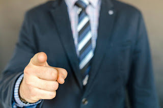 Image shows a masculine-presenting person, wearing a dark blue suit, a light blue shirt and a tie with diagonal stripes in differing shades of blue, pointing a finger towards the camera.