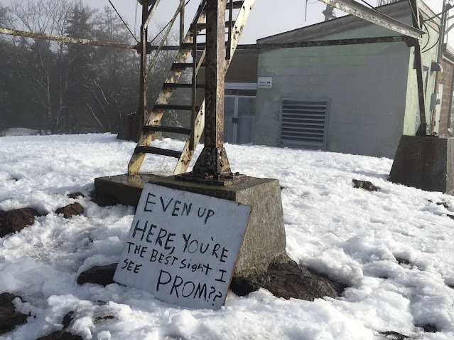 Hand-painted sign in the snow at the base of a tower staircase reads "Even up here you're the best sight I see. Prom?"