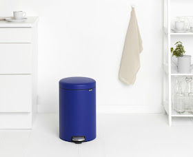 Royal blue pedal bin sitting in what is probably a bathroom; a towel hangs from a hook on the wall. The room is decoarted all in whites and off-whites so the bin really stands out.