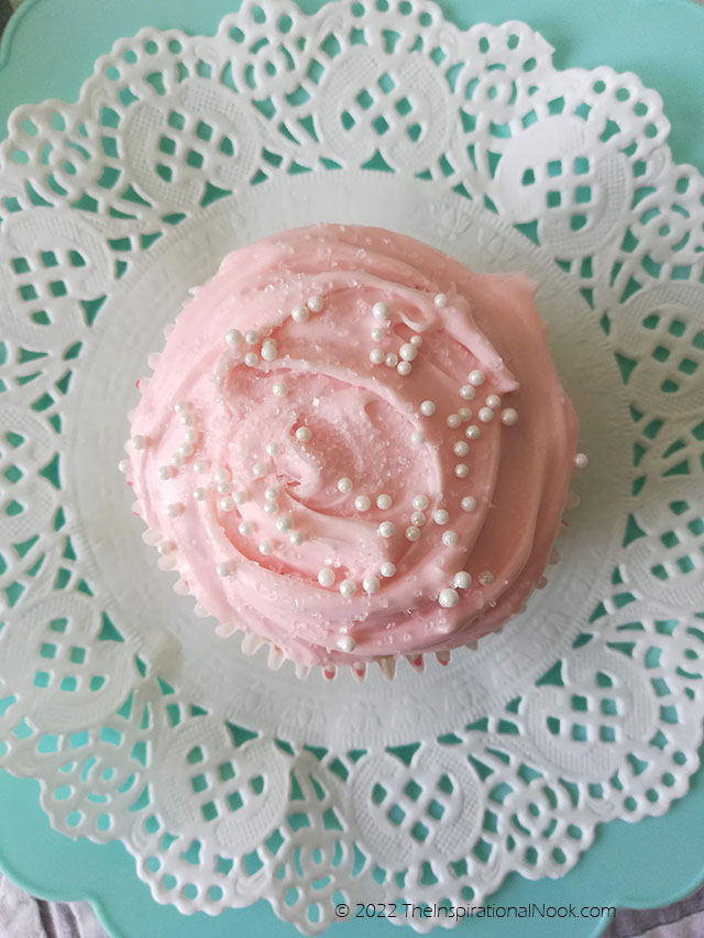 Pink birthday cupcake with pink frosting and white pearl non-pareils on a paper doily on a turquoise plate, Mother's Day cupcake