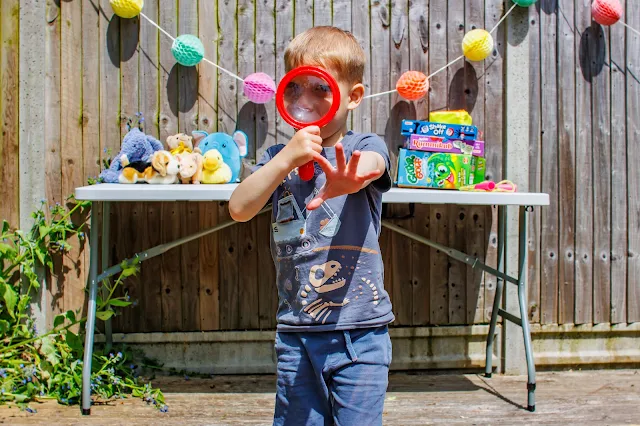 A child looking through a large magnifying glass with lots of toys on a table behind